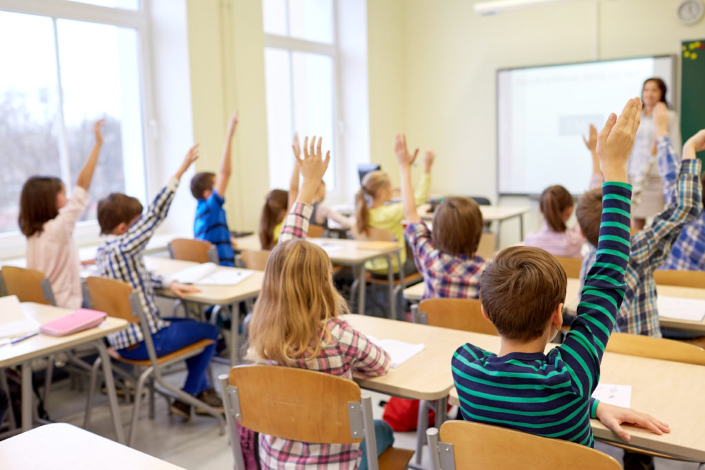 group of school kids raising hands in classroom